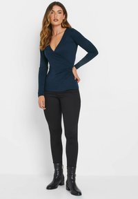Marine langærmet top med en dyb V-udskæring og intrikat indpakningsdesign, parret med sorte leggings og ankelstøvler. Glat stofstruktur.