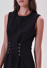 Frau in einem ärmellosen schwarzen Kleid mit einem Korsett-ähnlichen Schnürbund und einer massiven goldenen Kettenhalskette.