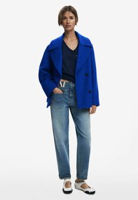 Manteau double boutonnage texturé bleu, haut marine, jean décontracté bleu clair, et chaussures noir et blanc. Design simple avec des accents de boutons remarquables.