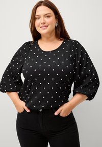 Zwarte blouse met witte stippen, gestructureerde stof, ronde halslijn en pofmouwen; gecombineerd met zwarte skinny jeans.