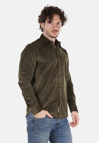 Camicia a maniche lunghe verde oliva realizzata in tessuto morbido, con chiusura con bottoni sul davanti, colletto classico e texture sottile. Abbinata a jeans blu.