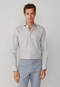 Weißes Langarmhemd mit dünnen grauen vertikalen Streifen, Kentkragen und kontrastierenden gestreiften Manschetten, kombiniert mit hellblauen Hosen.