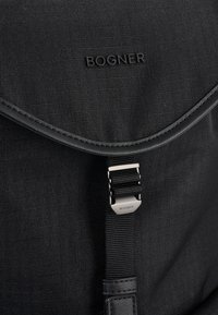 Črn nahrbtnik iz tkanine z teksturirano površino, z ukrivljenim pokrovom, metalno sponko in vtisnjenim napisom "BOGNER".