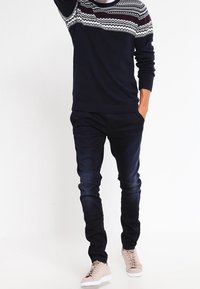 Marinblå tröja med vit och vinröd mönstrad yoke, parat med mörka jeans och ljusbeige sneakers. Åtsittande design, slät textur.