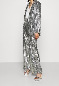 Stříbrný flitrový blazer a odpovídající kalhoty, které mají volný střih, dlouhé rukávy a hladkou texturu. V kombinaci s metalickými platformovými podpatky.