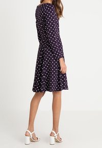 Kvinna klädd i en knälång, långärmad marinblå klänning med vita prickar, i kombination med vita högklackade sandaler, stående på sidan.