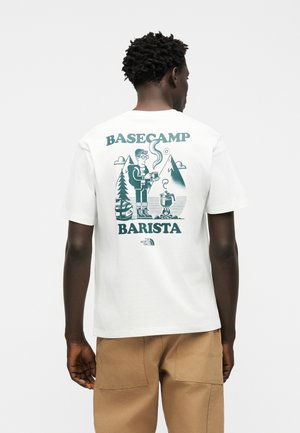 Mand iført hvid T-shirt med grønt "Basecamp Barista"-motiv, der viser en person, der laver kaffe udendørs med bjerge og træer.