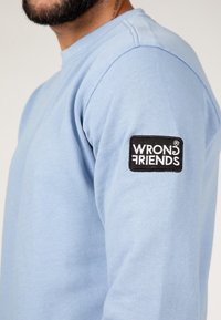 Hombre con una camisa de manga larga azul claro, con un parche en blanco y negro que dice "WRONG FRIENDS" en la parte superior del brazo izquierdo visible desde el lado.