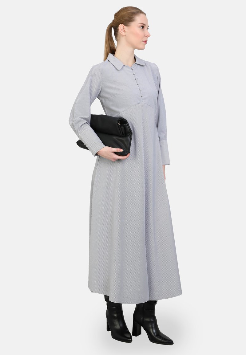 Pitkä, vaaleanharmaa mekko, jossa on kaulus ja edessä nappikiinnitys; sisältää pitkät hihat ja tekstuuripintaa. Musta clutch-laukku ja korkokengät.