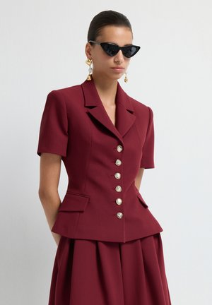 Femme portant une veste ajustée bourgogne à manches courtes avec des boutons dorés, une jupe assortie, des lunettes de soleil noires en forme de chat et des boucles d'oreilles pendantes dorées.