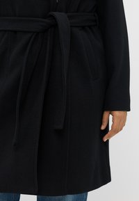 Zwarte wollen blend jas met een verstelbare taille, lange mouwen en zichtbare zijzakken. Glad textuur en klassiek ontwerp.