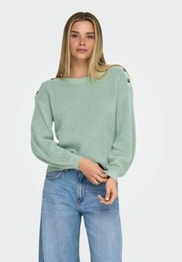 Femme aux longs cheveux blonds portant un pull en tricot vert clair avec des détails de boutons sur les épaules et un jean bleu clair.