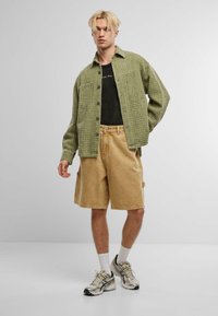 Chaqueta texturizada verde oliva sobre una camiseta de malla negra, combinada con pantalones cortos color beige y zapatillas gris. Cuenta con botones visibles y bolsillos laterales.