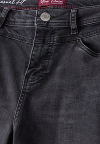Jeans de mezclilla negra con un corte informal, con costuras en contraste, cierre de botón metálico y cuatro bolsillos. Se aprecia una textura vintage.