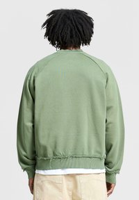 Felpa verde con design raglan, caratterizzata da un orlo sfrangiato, tessuto testurizzato e polsini a costine. Nessun motivo o dettaglio visibile.