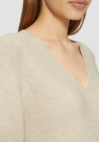 Jasno beżowy sweter z dzianiny z dekoltem w serek, teksturowanym materiałem i luźnym krojem z delikatnymi poziomymi paskami na powierzchni.