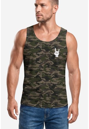 Mann mit kurzen Haaren, der ein grün-braunes Tarn-Top mit einem weißen Handzeichen und blaue Jeans trägt und vor einem schlichten Hintergrund steht.