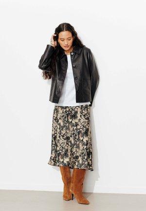 Femme aux longs cheveux foncés portant une veste en cuir noire, une chemise blanche, une jupe à motifs et des bottes hautes en daim marron, debout contre un mur blanc.