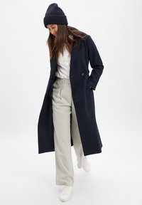 Marineblauer langer Mantel aus einer Wolle-Mischung, ausgestattet mit einem doppelreihigen Knopfdesign. Kombiniert mit hellgrauen, hoch taillierten Hosen und weißen Turnschuhen.