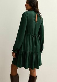 Kobieta w ciemnozielonej, długiej sukience z teksturą, z dekoltem na plecach i paskiem w talii, w ciemnych kozakach sięgających do kolan, stojąca na tle jasnej ściany.