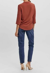 Rostfärgad topp med trekvartsärmar, kombinerad med blå denimjeans och beige högklackade skor. Slät textur, avslappnad passform, rundad nederkant.