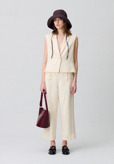 Un gilet créme et un pantalon large en tissu léger. Un chapeau seau marron foncé, des mocassins noirs et un sac à main bordeaux complètent le look.