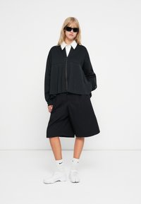Jaqueta preta curta com detalhes em pregas, combinada com calções pretos largos. Uma camisa branca com colarinho, sapatos de desporto brancos e óculos de sol completam o visual.