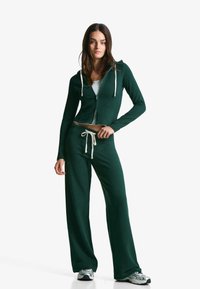 Mørkegrøn zip-up hoodie med hvide snore, parret med matchende brede bukser. Materialet ser blødt og struktureret ud, velegnet til afslappet brug.
