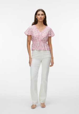 Vrouw staand, gekleed in een roze bloemen V-hals blouse met zwierige mouwen, witte broek met hoge taille, en sandalen met bandjes, tegen een effen achtergrond.