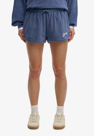 Osoba ubrana w niebieskie spodenki z frotty ze sznurkiem, białe skarpetki i beżowe trampki, stojąca na jednolitym białym tle.