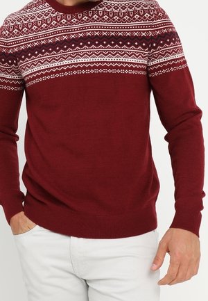 Mann trägt einen kastanienroten Pullover mit weißen geometrischen Mustern über Brust und Schultern, kombiniert mit hellen Hosen.