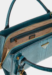 Borsa a mano in turchese con una texture liscia e lucida. Caratterizzata da un logo dorato, scomparti con cerniera e manici robusti. Interno foderato.