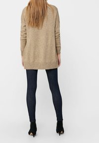 Nainen seisoo selkä kameraan päin, päällään pitkä beige neule, tummat skinny farkut ja mustat korkeat nilkkurit yksinkertaista valkoista taustaa vasten.