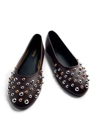 Zapatos planos marrones con forma redondeada, adornados con tachuelas metálicas en plata, oro y bronce, sobre una textura lisa similar al cuero.