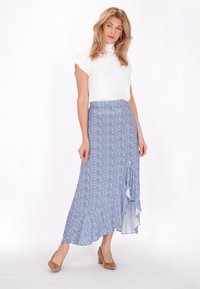 Weißes kurzärmliges Oberteil, kombiniert mit einem langen, blauen, blumigen Rock mit einem gerafften Saum und einem hohen Schlitz, ergänzt durch beige High Heels.