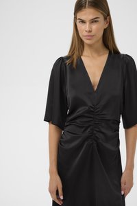 Melna kleita ar īsām pūkainām piedurknēm, dziļu V-veida izgriezumu un savilkumu detaļām viduklī un svārkos, izgatavota no gludas, teksturētas auduma.