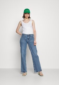 Top de alças branco, jeans largos azuis, boné verde com o texto "LAS PALMAS" e sandálias strappy verde claro. Conjunto simples e casual.