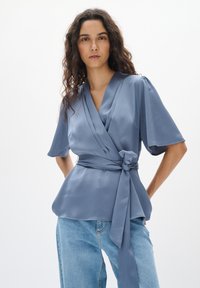 Blå texturerad wrap-blus med v-ringning och korta ärmar, knuten i midjan med ett matchande bälte. Kombinerad med ljusblå denimjeans.