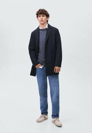 Joven con cabello rizado que lleva puesto un abrigo negro, suéter gris, camisa blanca, jeans azules y zapatillas beige de pie contra un fondo liso.
