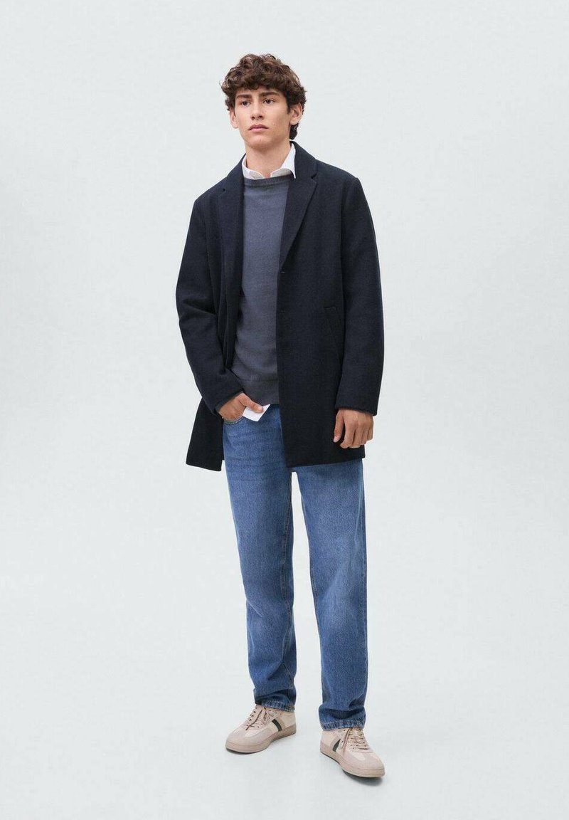 Joven con cabello rizado que lleva puesto un abrigo negro, suéter gris, camisa blanca, jeans azules y zapatillas beige de pie contra un fondo liso.