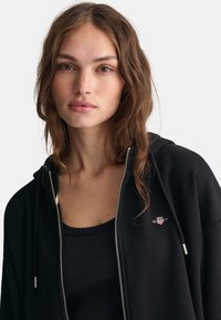 Sort zip-up hættejakke med sølvdetaljer og logo-accent, parret med en tætsiddende sort tanktop, begge i et glat, blødt stof.