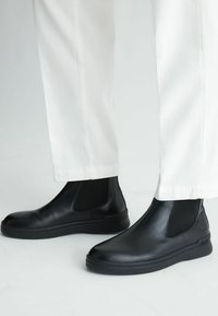 Stivali Chelsea neri in pelle con pannelli elastici laterali, texture liscia, punta arrotondata e suola in gomma spessa, abbinati a pantaloni bianchi.