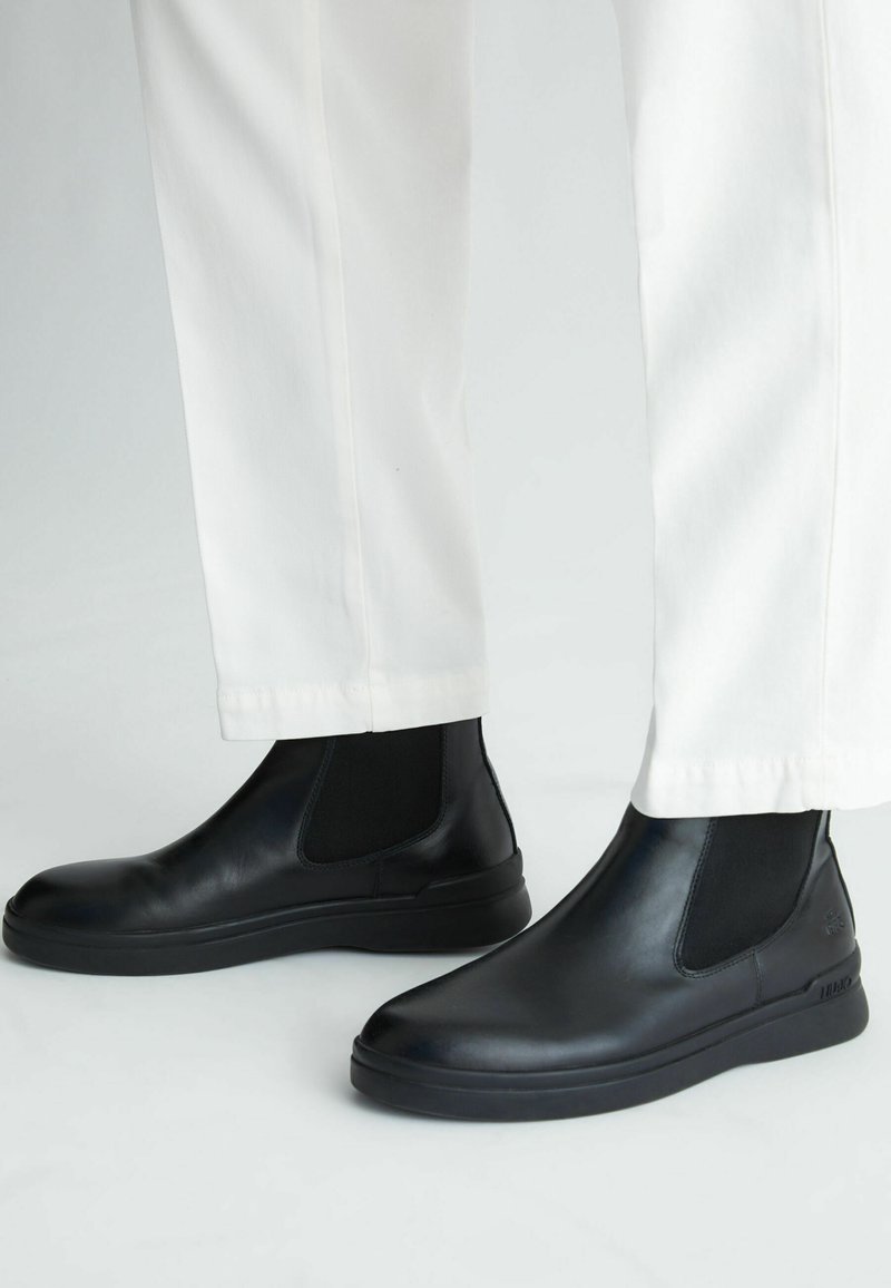 Stivali Chelsea neri in pelle con pannelli elastici laterali, texture liscia, punta arrotondata e suola in gomma spessa, abbinati a pantaloni bianchi.