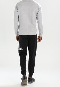 Sweatshirt gris avec manches raglan et pantalon de jogging noir présentant un logo blanc sur la cuisse gauche. Des chaussures de sport noires sont portées.