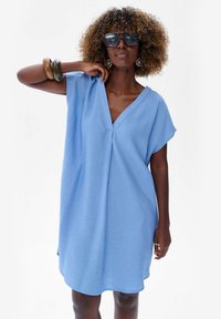 Jasnoniebieska sukienka z krótkim rękawem, głębokim dekoltem w kształcie V, luźnym kroju i miękkiej teksturze. Posiada subtelne detale w przeszyciach oraz rozcięcia boczne.