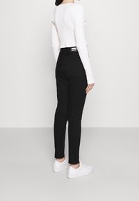 Jeans de mezclilla negra con cintura alta y corte ajustado, que presentan un parche de logo en la parte trasera y dobladillos enrollados, combinados con zapatillas blancas.