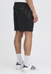 Svarta shorts med en slät textur, elastisk midja och två fickor med dragkedja. Bärs med vita strumpor och svartsrandiga sneakers.