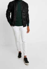Uomo che indossa una giacca di pelle nera, un maglione verde scuro, pantaloni bianchi e sneakers nere, mentre cammina su uno sfondo bianco uniforme.