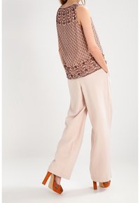 Bezramienna koszulka w wzory z jasnobrązowym geometrycznym wzorem, zestawiona z luźnymi beżowymi spodniami i pomarańczowymi butami na obcasie.