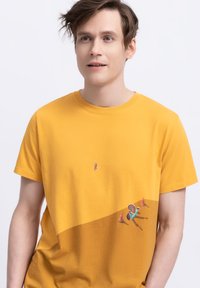 Geel katoenen T-shirt met ronde hals, met een kleine afbeelding van een persoon en objecten in de rechter onderhoek, wat een contrast creëert met de stof.
